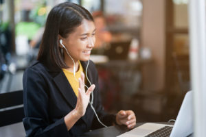 A lady wears earphones and smart clothes while on skype. She is waving at the camera greeting colleagues.