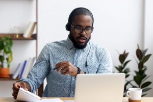 A man takes part in a remote interview at his laptop. He has on a headset and is holding some notes.