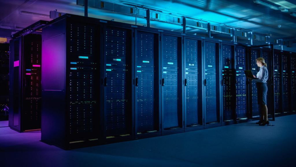 A woman looks down at her laptop standing in front of data servers, in our article we look to the world of edge computing and the differences between micro data centres and regional data farms
